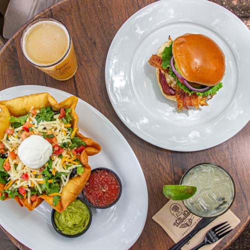 A burger with bacon and lettuce, a salad in a tortilla bowl, and drinks on a wooden table.