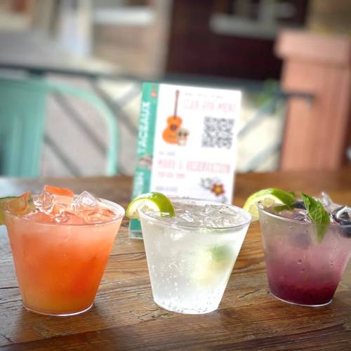 Three colorful cocktails on a wooden table, featuring a red drink, a clear drink, and a purple drink, garnished with lime.