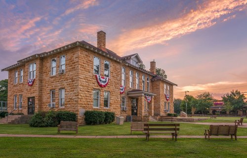 Historic stone building adorned with patriotic bunting, set against a colorful sunset sky and green lawn.