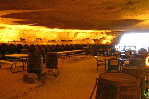 A dimly lit cave interior with wooden barrels, tables, and chairs arranged on a sandy floor.