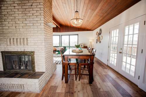 Cozy dining area with a wooden table, chairs, and a brick fireplace, featuring large windows and a warm wooden ceiling.