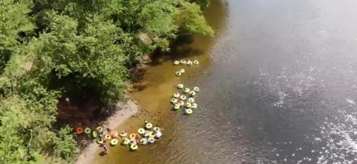 Aerial view of a riverbank with people floating on colorful inner tubes among trees and calm water.