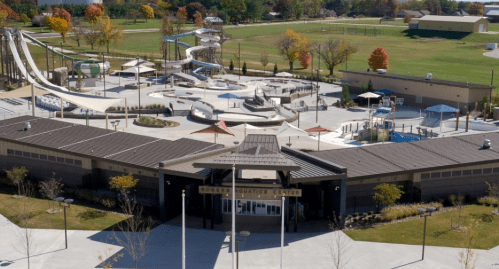 Aerial view of a modern aquatics center with water slides, pools, and landscaped areas in a park setting.