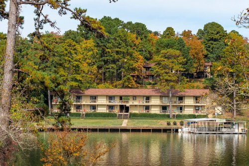 A lakeside view of a multi-story building surrounded by trees with autumn foliage, reflecting in the water.