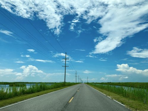 A long, straight road flanked by water and tall grass under a bright blue sky with scattered clouds.