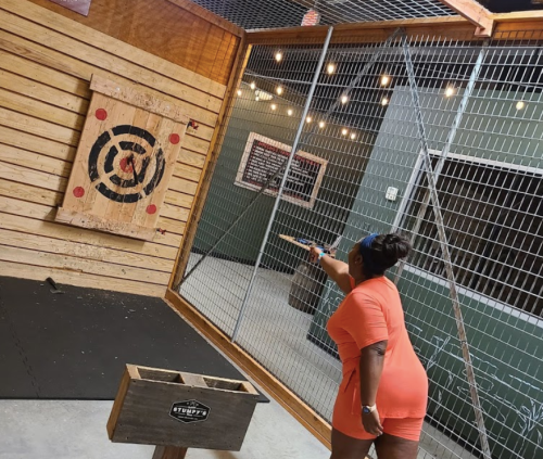 A person in an orange outfit throws an axe at a target in an indoor axe-throwing venue.
