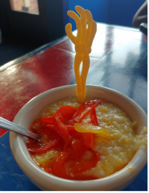 A bowl of creamy porridge topped with sliced red peppers and a quirky yellow hand-shaped stirrer.