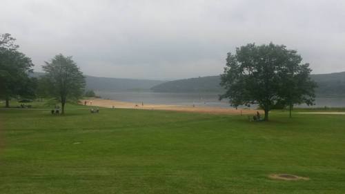 A serene lakeside view with a sandy beach, grassy areas, and trees under a cloudy sky. People are enjoying the outdoors.