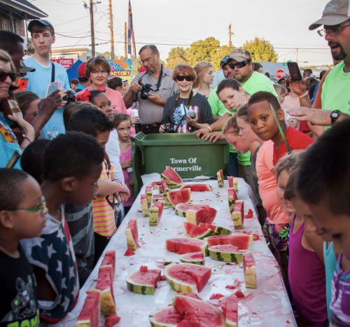 A lively crowd of children and adults gathers around a table filled with watermelon slices at a community event.