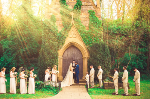 A couple exchanges vows in front of a charming chapel, surrounded by wedding party members in a lush, green setting.