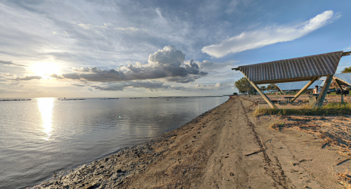 A serene beach scene at sunset, with calm waters, clouds, and a small shelter on the shore.
