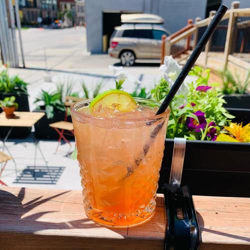 A refreshing cocktail with ice and a lime slice, set on a wooden railing with plants and a car in the background.