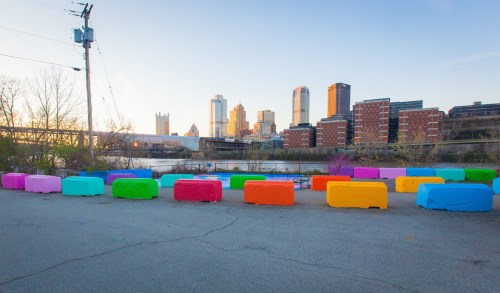 Colorful painted blocks line a waterfront area with a city skyline in the background during sunset.