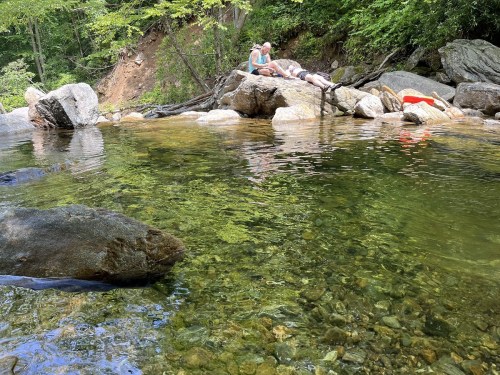 A serene river scene with clear water, rocks, and two people relaxing on a large rock by the water's edge.