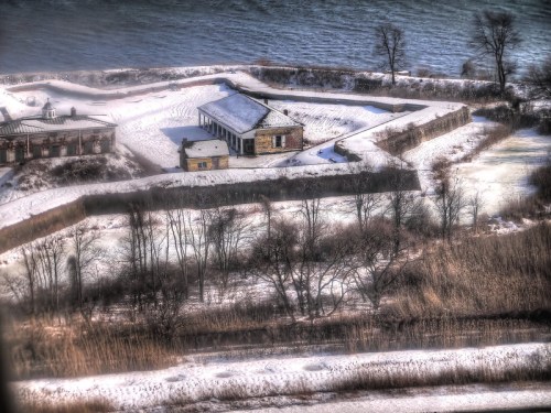 A snowy landscape featuring a historic building surrounded by trees and water in the background.