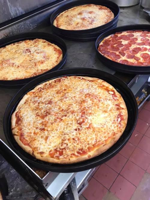 Four pizzas in black pans, including a cheese pizza and a pepperoni pizza, on a baking surface.