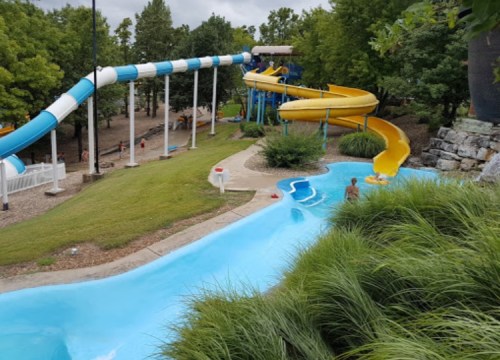 A water park scene featuring two colorful water slides and a lazy river surrounded by greenery and trees.