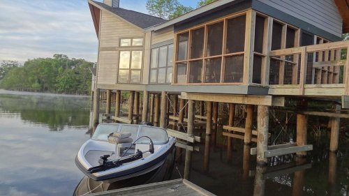 A boat docked beside a modern house on stilts, overlooking a calm river surrounded by trees.