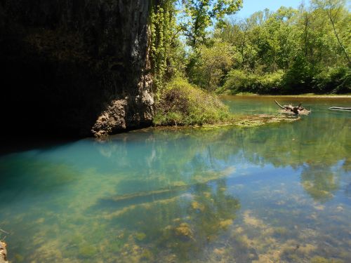 A serene river scene with clear blue water, lush greenery, and rocky cliffs under a bright blue sky.
