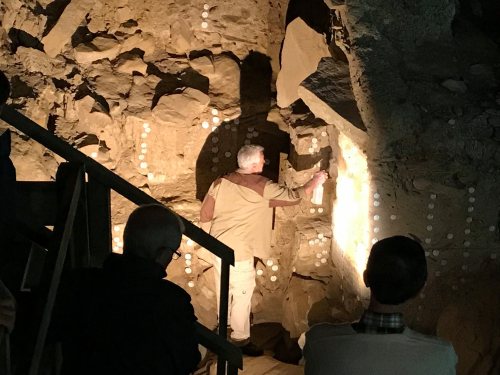 A person examines rock formations in a dimly lit cave, with others observing from a distance.