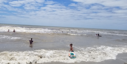 A child with a float walks along the shore, while others swim in the ocean under a cloudy sky.