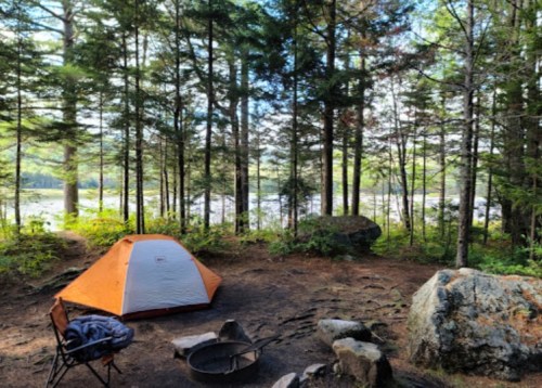 A cozy orange and white tent set in a forest clearing by a lake, surrounded by trees and a fire pit nearby.