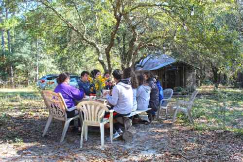 A group of people gathered around a table outdoors, surrounded by trees and a rustic shed in the background.