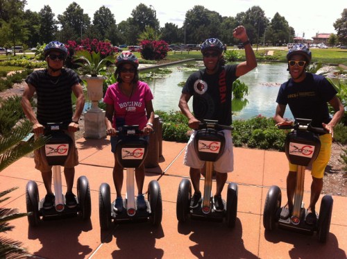 Four people on Segways, smiling and posing near a pond with flowers in a sunny park.