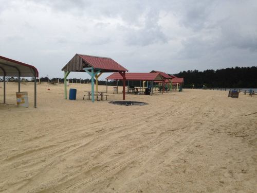 Beach scene with several picnic pavilions, sandy ground, and a cloudy sky in the background.