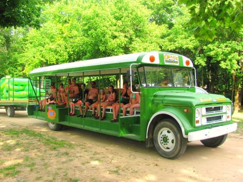 A green bus with passengers sitting inside, surrounded by trees and a trailer with inflatable water toys.