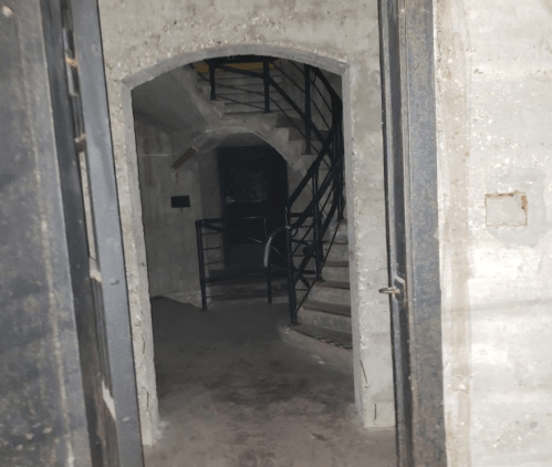 A dimly lit concrete hallway with a staircase and metal railing, leading into a darkened area.