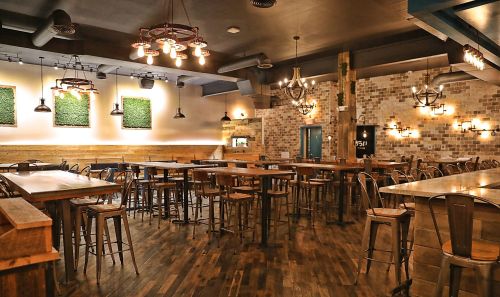 A spacious restaurant interior with wooden tables, metal chairs, and decorative lighting. Green wall art adds a touch of nature.