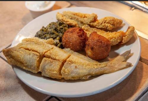 A plate of fried fish, cornbread balls, and greens, served with a side of dipping sauce.