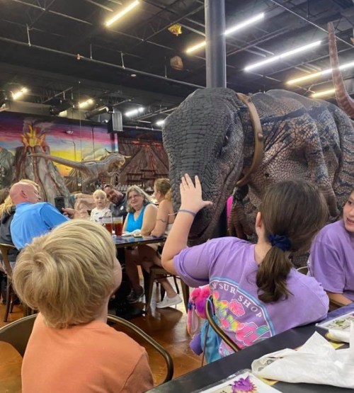 A child reaches out to a life-sized dinosaur model in a restaurant, while families enjoy their meals in the background.