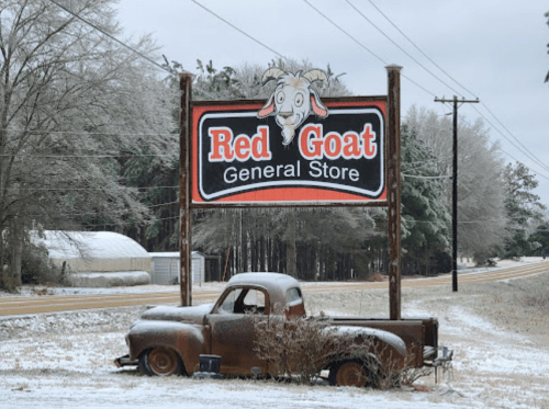 A vintage truck parked near a sign for Red Goat General Store, surrounded by icy trees and a rural road.