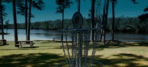 A disc golf basket near a calm lake, surrounded by trees and picnic tables under a clear blue sky.