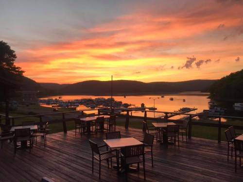 A vibrant sunset over a lake, with boats docked and empty tables on a wooden deck in the foreground.