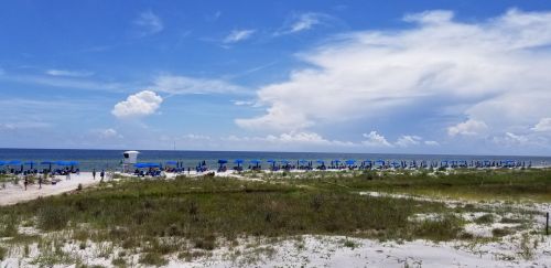 A sunny beach scene with blue umbrellas, white sand, and a calm ocean under a partly cloudy sky.