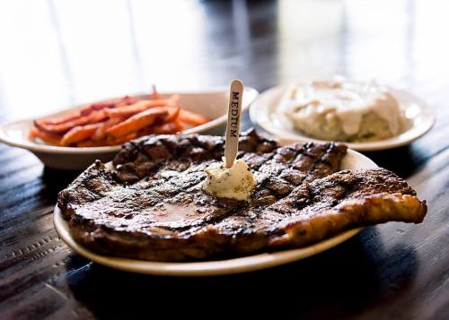 A large grilled steak with a dollop of butter, served with sweet potato fries and a side of creamy mashed potatoes.