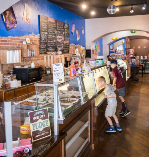 Two children explore a colorful ice cream shop, peering into display cases filled with treats. Bright decor and shelves are visible.