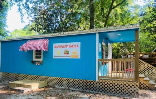 A bright blue food stand with a striped awning, featuring a sign that reads "Outpost Grill" and a wooden deck.