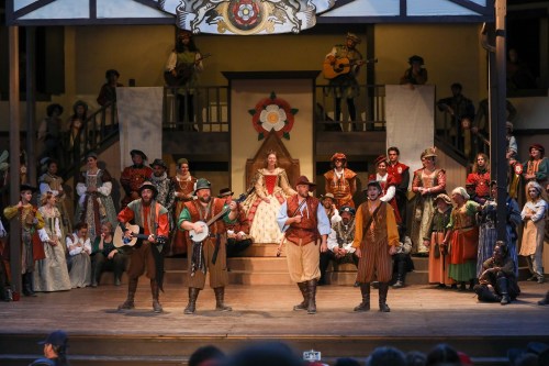 A lively theater performance with actors in colorful costumes, musicians, and a central figure in a white dress on stage.