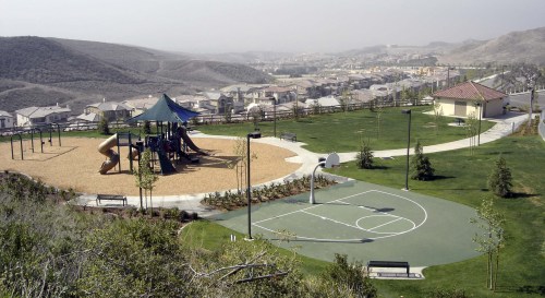 A playground with a slide and swings next to a basketball court, overlooking a residential area and hills.