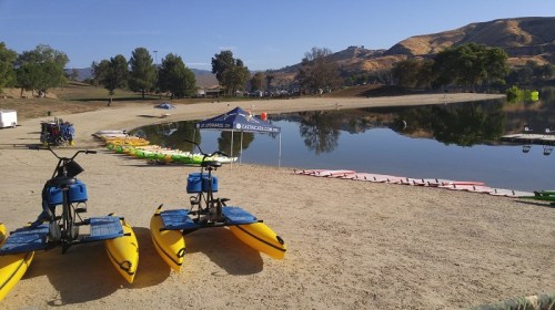 Colorful kayaks and paddleboards lined up on a sandy shore by a calm lake, with trees and hills in the background.