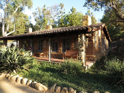 A rustic stone cottage with a tiled roof, surrounded by greenery and trees, featuring a wooden bench on the porch.