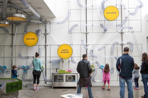 Visitors interact with a colorful wall installation featuring pipes and city names, engaging in a hands-on exhibit.