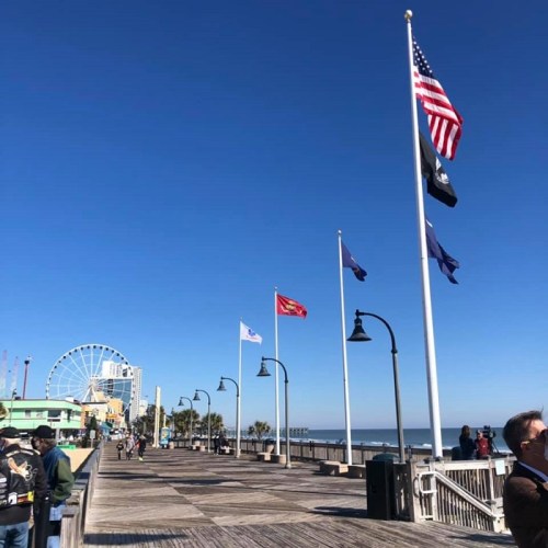 A sunny boardwalk with flags, a ferris wheel in the background, and people enjoying the day by the ocean.