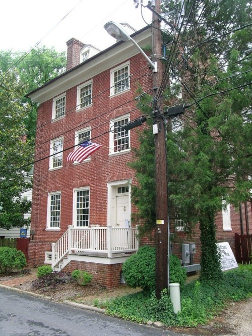 A three-story brick house with a front porch, an American flag, and surrounding greenery on a quiet street.