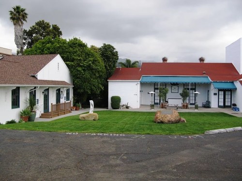 Two buildings with red roofs and white walls, surrounded by green grass and trees, under a cloudy sky.