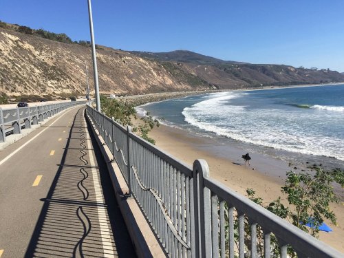 A scenic coastal view with a bike path alongside the beach, waves crashing, and a person walking on the sand.